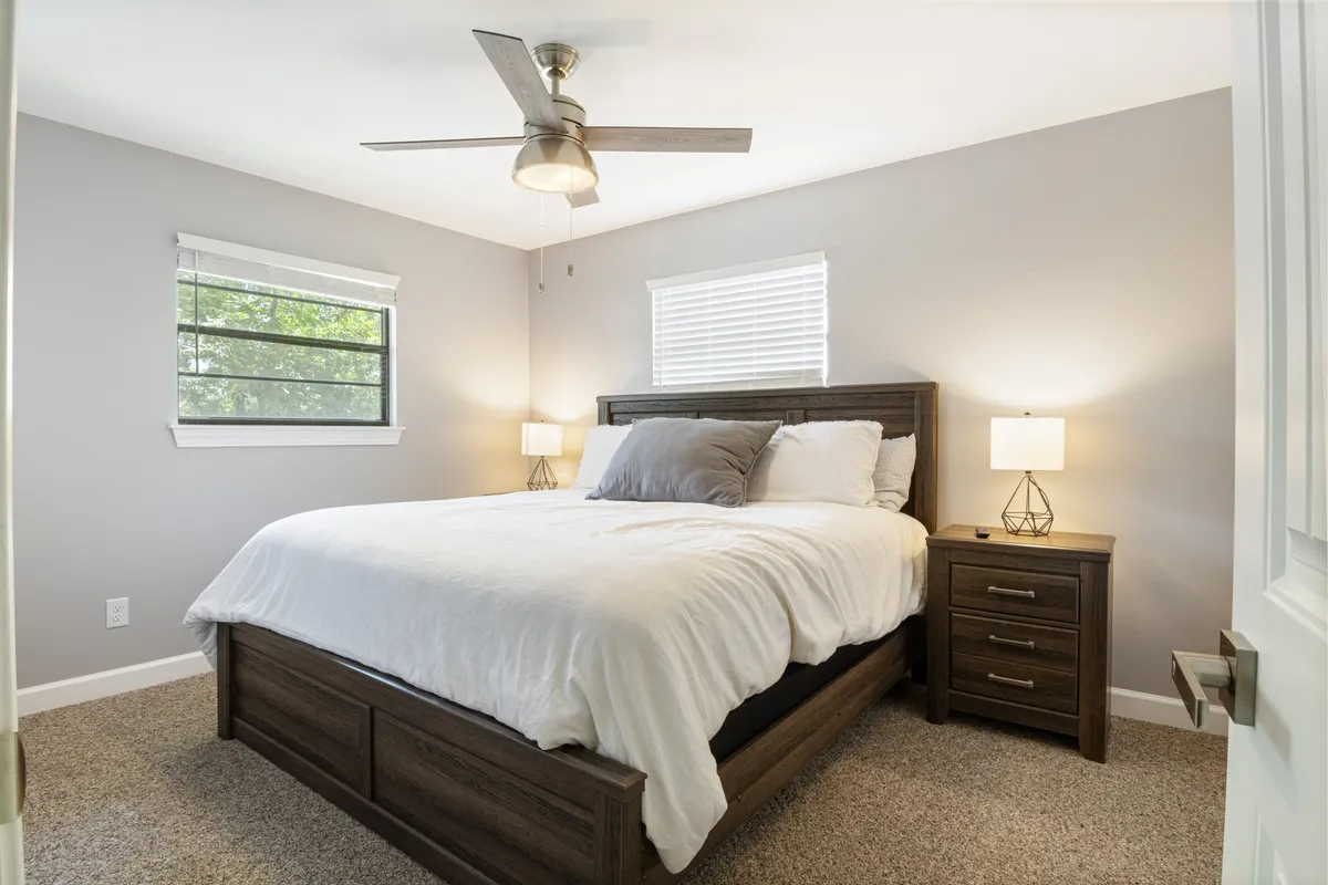 This image features a serene bedroom with a plush bed, nightstand, soft lighting from lamps, and large windows letting in refreshing morning light. The decor exudes calm and tranquility, perfect for a peaceful night's sleep or reading in bed.