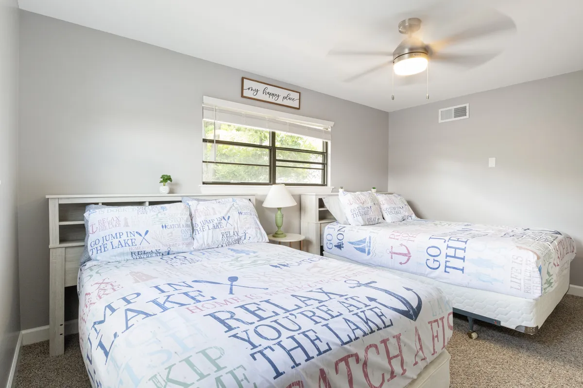 This image showcases a bedroom with two beds featuring decorative bedding, plenty of natural light pouring in from the window, a cozy nightstand with a lamp, and a decorative wall sign. The space feels spacious, tidy, and perfect for relaxation or reading.