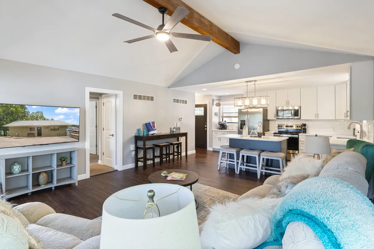A bright, open-concept living and dining area with a large dining table ready for shared meals, a kitchen island with bar stools for casual dining, a cozy sofa with colorful pillows, and a television for entertainment. The space is well-lit and inviting, supporting both dining and social gatherings.