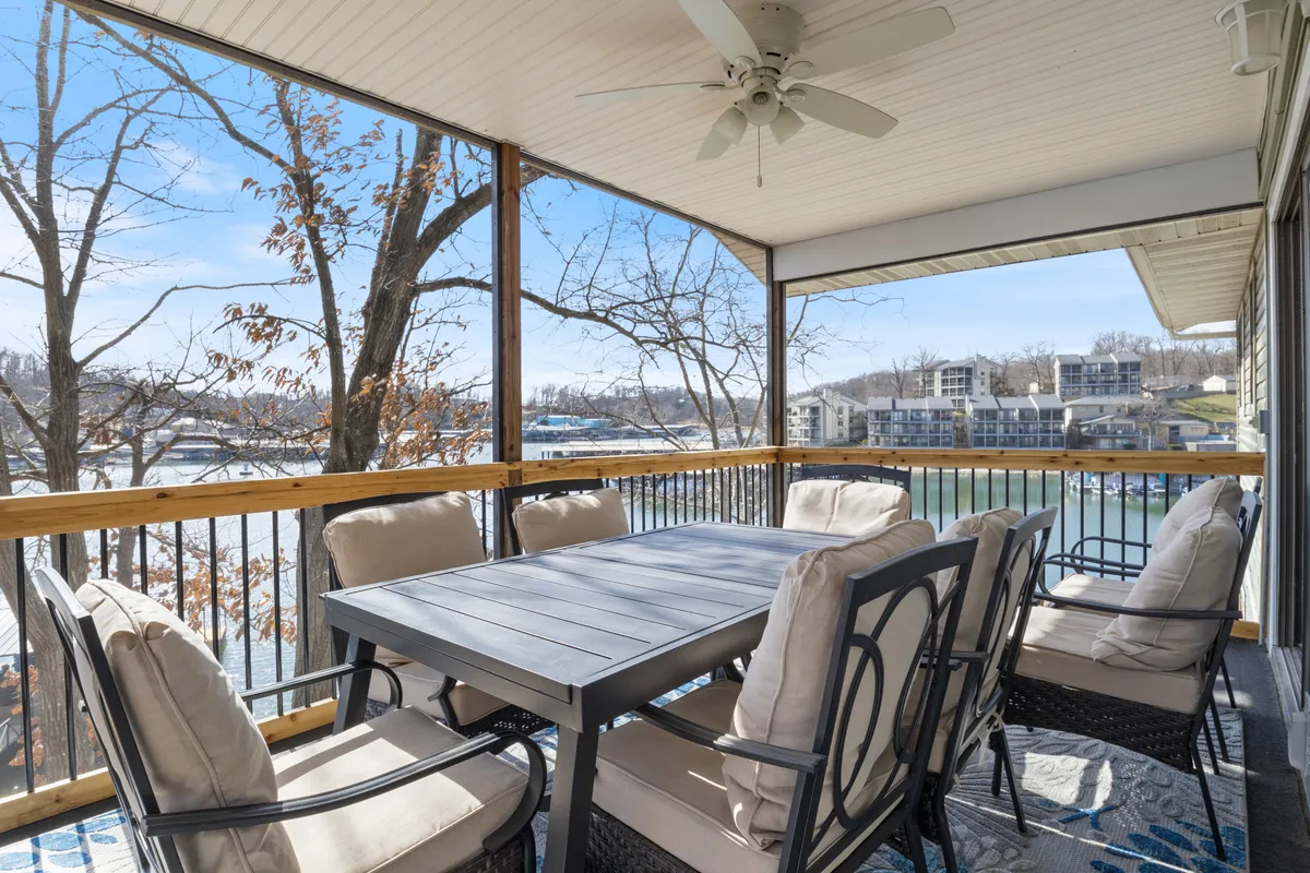 An inviting outdoor dining area with a large table and comfortable seating, all overlooking a tranquil body of water. Surrounded by lush trees, the space exudes a serene, relaxed vibe, and hints at group gatherings, meals, and sunset conversations.