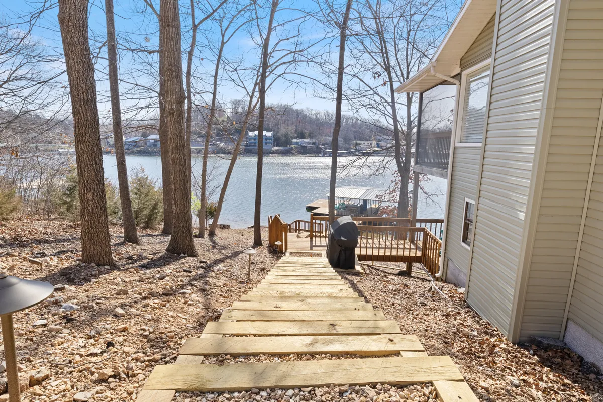 A wooden staircase leads down to a spacious deck area overlooking the lake, framed by trees. The deck is open, with plenty of space for activities like cornhole, stargazing, or simply taking in the sunrise over the water.