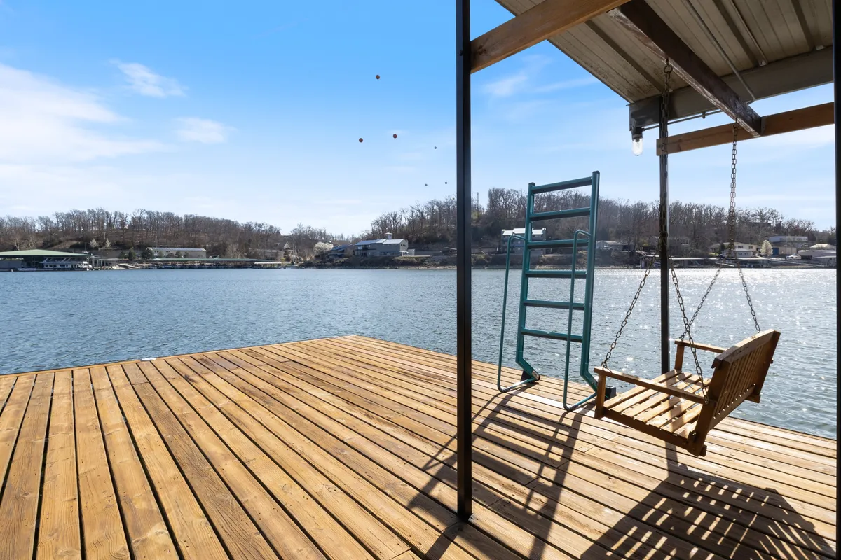 The image showcases a tranquil lakeside scene featuring a wooden dock extending over calm waters, a cozy swing perfect for relaxing, a ladder leading into the lake, and picturesque hills in the background. The setting is serene, inviting guests to unwind, fish, or enjoy lakeside sunsets.
