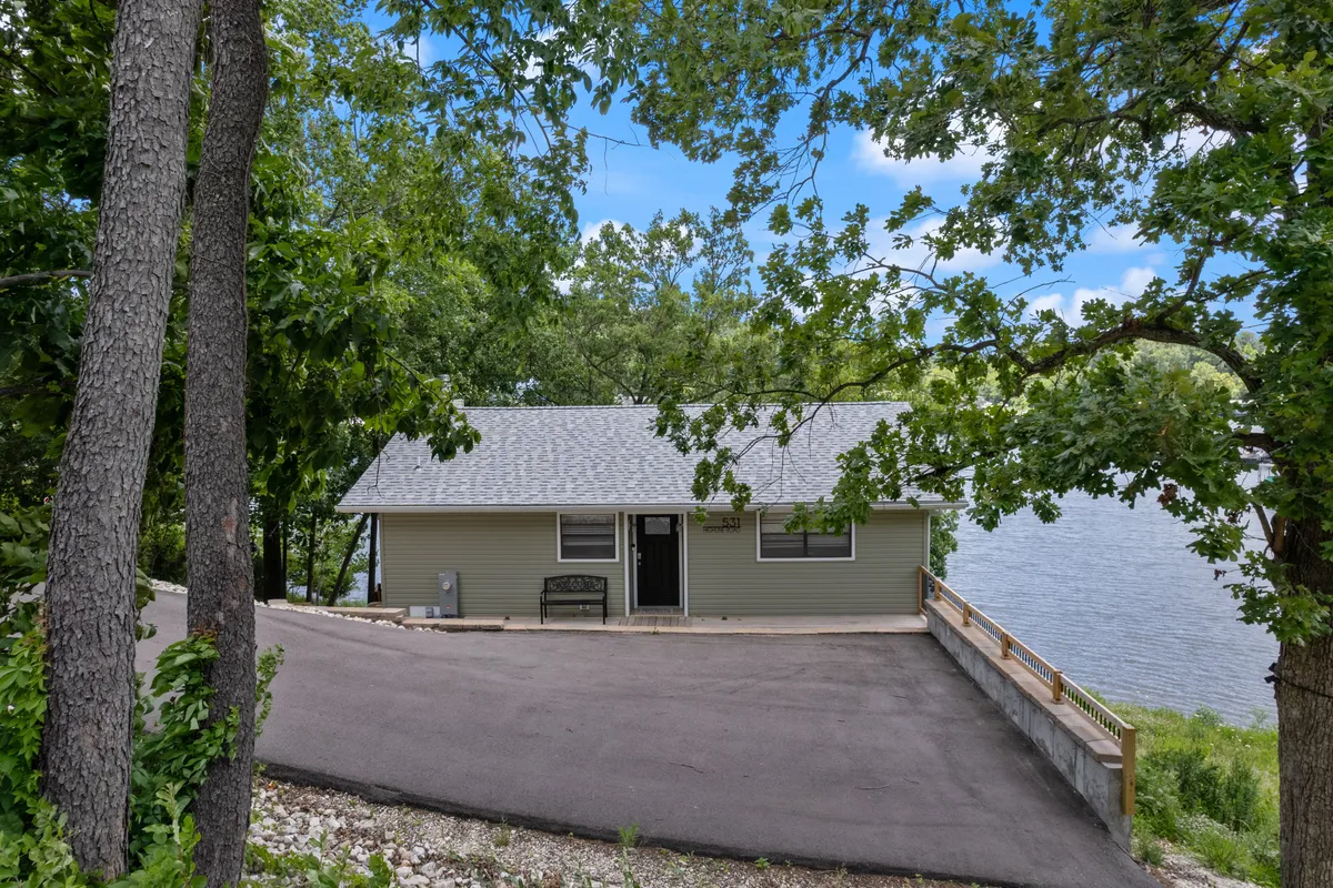 A welcoming exterior scene featuring the house, surrounding trees, driveway, and the waterway beyond. The setting sun casts a peaceful glow, suggesting both daytime fun and evening gatherings by the fire pit.