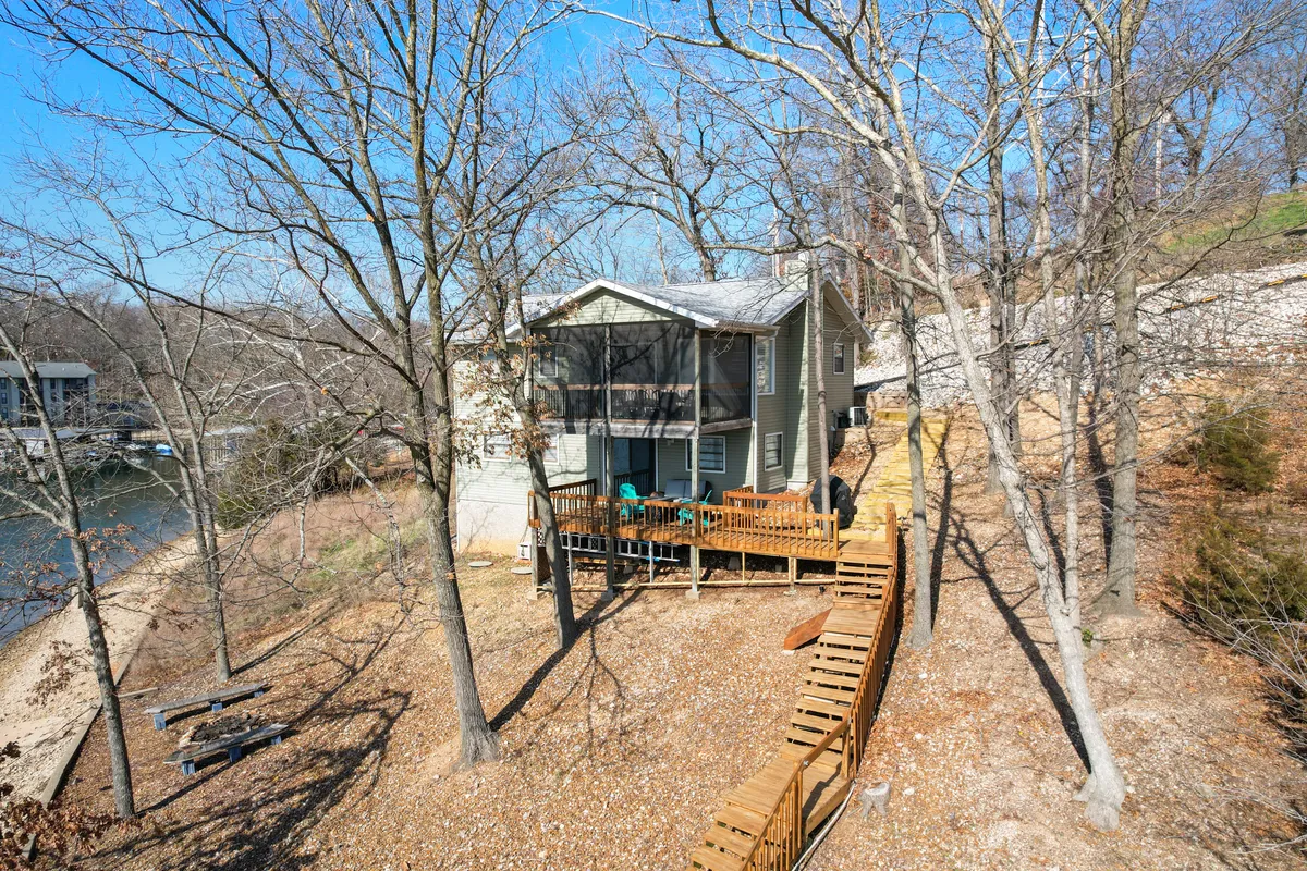 This image showcases the house with a welcoming porch, an inviting deck area, mature trees, and a clear pathway leading directly to the water’s edge. The tranquil setting highlights both the connection to nature and ease of outdoor access from the home.
