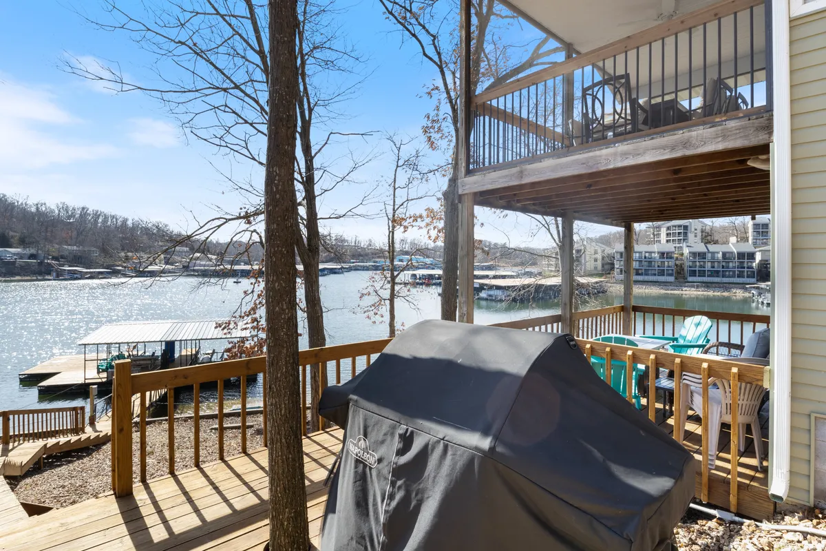 A waterfront deck with a grill and comfortable furniture set amid trees, offering an inviting space for barbecuing, relaxing, and socializing while enjoying views of the lake.