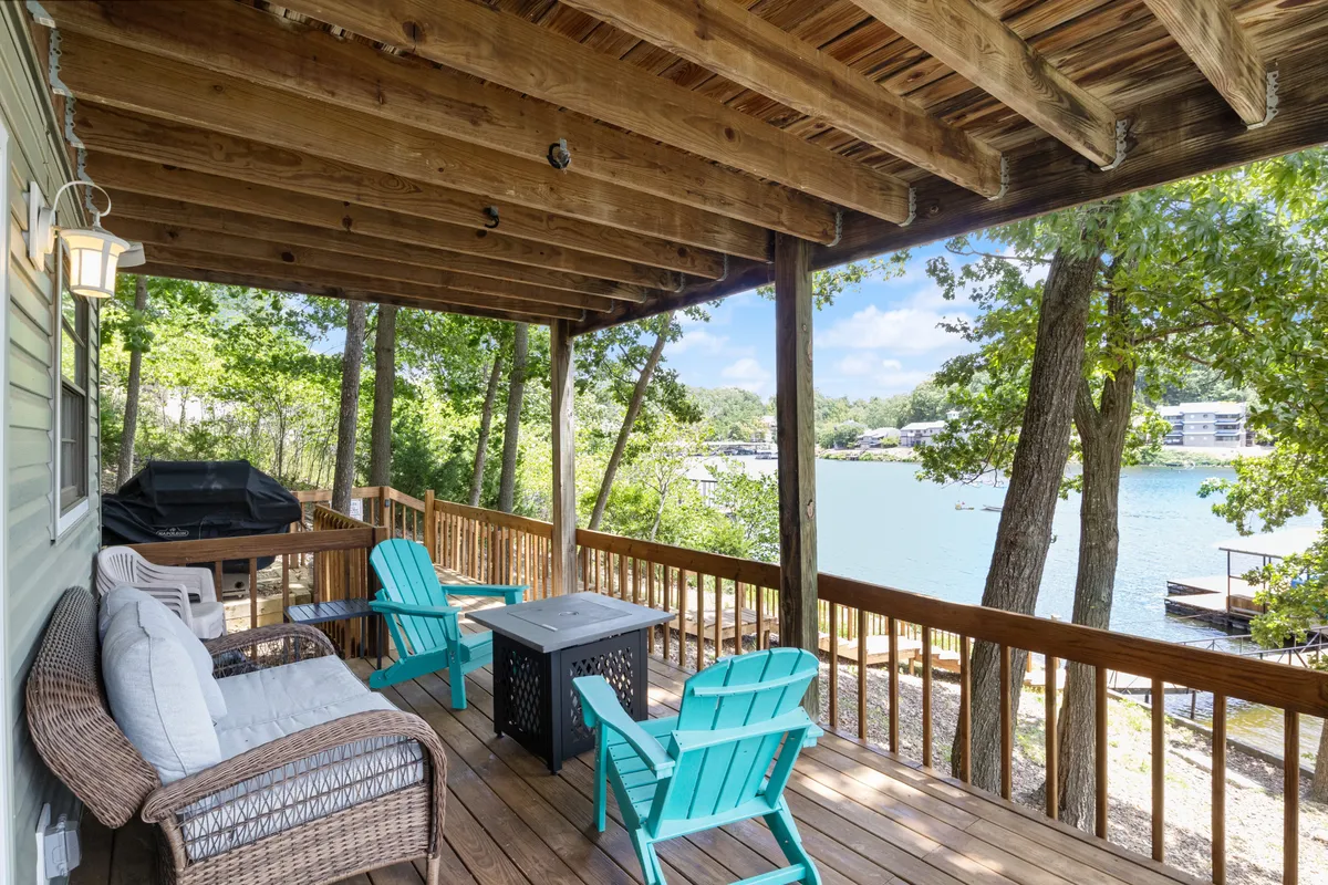 This image features a spacious wooden deck furnished with turquoise chairs and outdoor furniture, all overlooking a serene lake. The view emphasizes the seamless flow from the indoor living area to outdoor spaces, capturing the essence of lakeside living. The setup is perfect for lounging, dining, or soaking in the tranquil scenery.